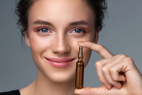 Woman holds ampoule with serum for hair or skin care. Photo of attractive woman with perfect makeup on gray background. Beauty concept